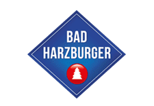 Das Logo von Bad Harzburger zeigt den Markennamen in wei&szlig;en Gro&szlig;buchstaben auf einem blauen, rautenf&ouml;rmigen Hintergrund. Unten im Logo befindet sich ein roter Kreis mit einer stilisierten wei&szlig;en Tanne.